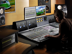 A person sits at a large professional audio mixing console with multiple monitors in a studio setting.