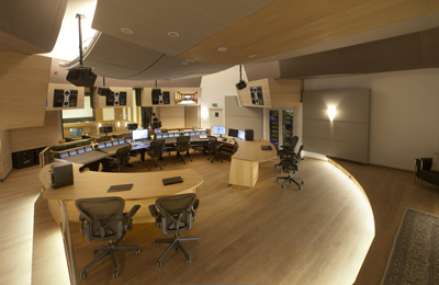 A wide view of a curved, modern control room with wooden desks, computer monitors, and ergonomic chairs.