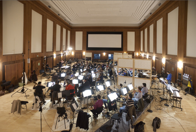 An orchestra sits in rows of chairs with music stands in a large, wood-paneled hall with a screen at the front.