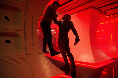 Two people in silhouette stand in a futuristic, red-lit curved hallway.