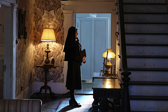 A nun in a black habit stands in a hallway next to a staircase and a lit lamp.