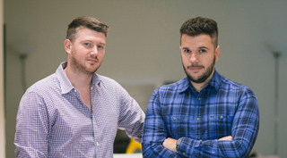 Two men posing indoors; one in a light checkered shirt and one in a blue plaid shirt with arms crossed.
