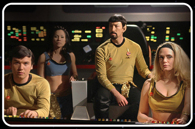 Four people in sci-fi costumes sit at control panels on a dark spaceship bridge.