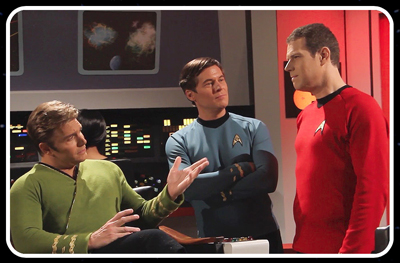 Three men in blue, red, and green Star Trek uniforms stand on a spaceship bridge.