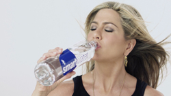 A blonde woman drinking water from a clear Smartwater bottle against a white background.