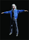 A person with long light hair wearing a blue jacket and dark pants stands with arms extended against black.