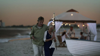 An older man and woman walk together on a sandy beach near a lit tent at twilight.