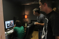 Three people stand and sit in a dimly lit room, observing a computer screen displaying an image.