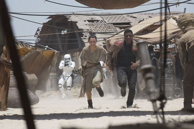 A woman and a man run through a dusty desert market while a stormtrooper chases them in the background.