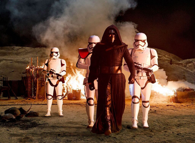 A hooded figure in black robes walks between two stormtroopers in front of a fire and smoke.