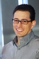 A smiling man with dark hair and glasses wearing a grey striped button-down shirt.