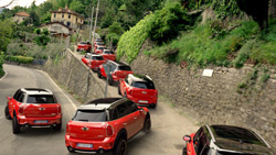 A line of red compact SUVs driving along a narrow, winding road next to a stone wall and lush green hillside.
