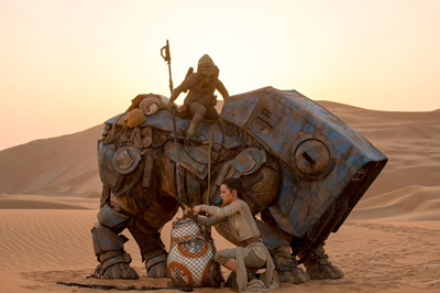 A person kneels by a large, blue mechanical creature in a desert at sunset, with another figure riding on top.
