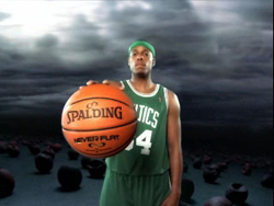 A basketball player in a green Celtics jersey holds a Spalding basketball against a dark, cloudy sky.