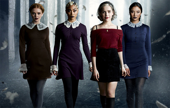 Four women in different colored dresses standing in a dark, misty forest.