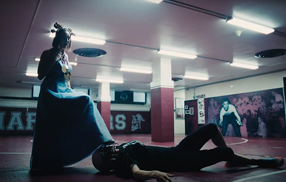 A woman in a long blue dress stands beside a man lying on the floor of a gymnasium.