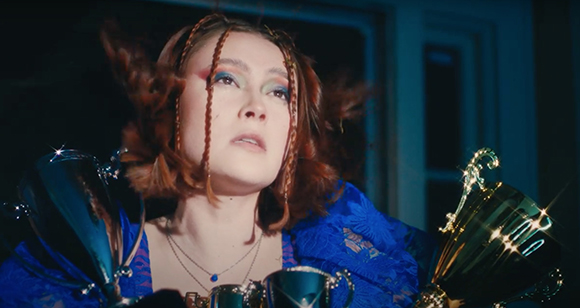 A person with reddish-brown braided hair and colorful eye makeup looks up while holding gold and silver trophies.
