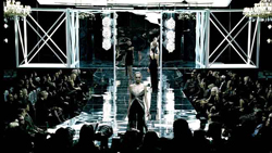 A model walks down a bright, reflective runway toward an audience in a dark room.