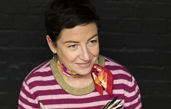 A woman with short dark hair and a patterned neck scarf smiles against a dark brick background.