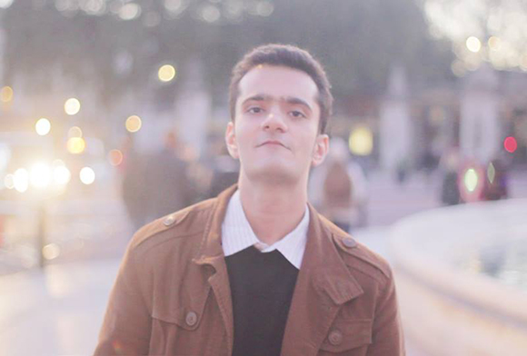 A young man in a brown jacket and black shirt stands in a blurred city square at dusk.