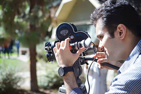A man looks through the viewfinder of a black professional film camera mounted on a tripod.