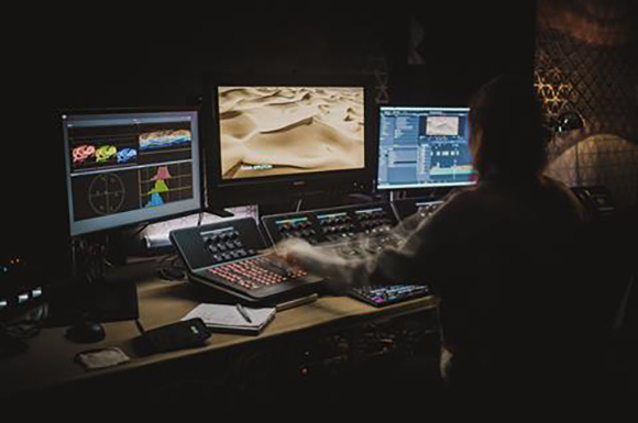 A person sits at a desk with three computer monitors and various control panels in a dark room.
