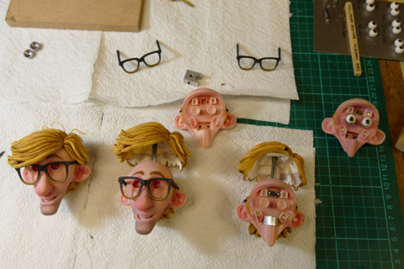 Several doll heads with blonde hair, some with glasses, scattered on white paper and a green cutting mat.