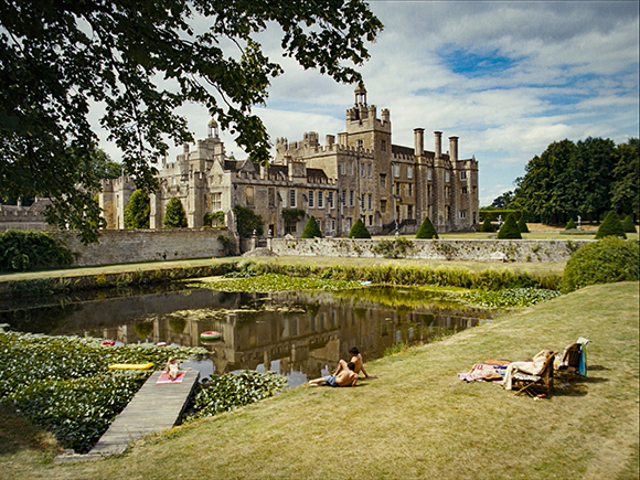 A large stone manor house reflected in a pond with people sunbathing on the grass nearby.