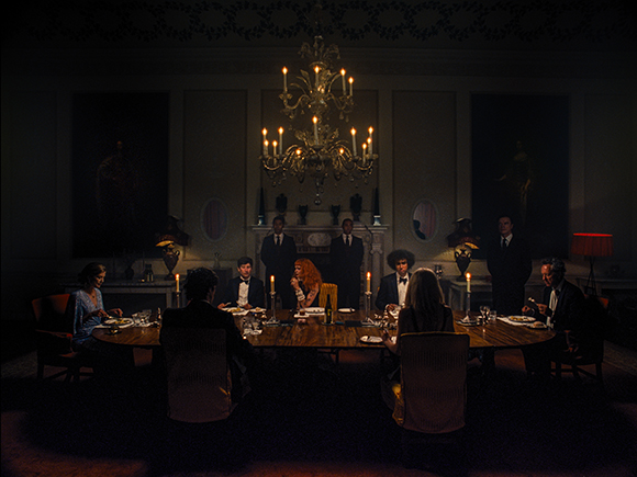 People seated around a long dining table in a dark, formal room lit by a large chandelier.