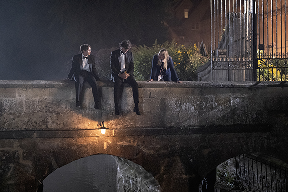 Three people in formal attire sit on the edge of a stone bridge at night.