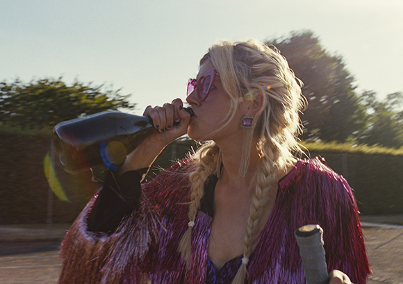 A blonde woman with braided hair drinks from a dark bottle while wearing pink sunglasses and a fringed jacket.