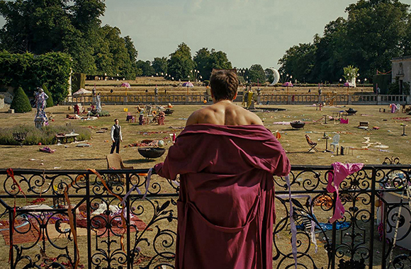 A person in a maroon robe stands at a balcony overlooking a vast field with scattered items and distant figures.