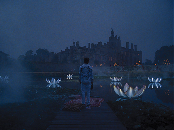 A person stands on a wooden dock facing a large castle across a dark lake filled with glowing lotus flowers.