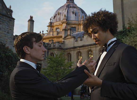 Two young men in black suits and bow ties hold hands in front of a large domed building.