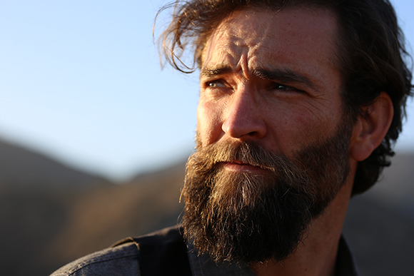 Close-up of a man with a thick brown beard and wavy hair looking off-camera during golden hour.