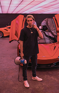 Man in black t-shirt and dark jeans holding a megaphone in front of an orange supercar with open doors.