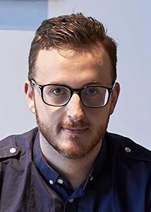 A man with short brown hair and a beard wearing black-rimmed glasses and a dark blue shirt.