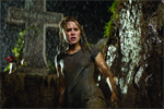 A woman with wet hair stands in heavy rain beside a large stone cross in a dark setting.