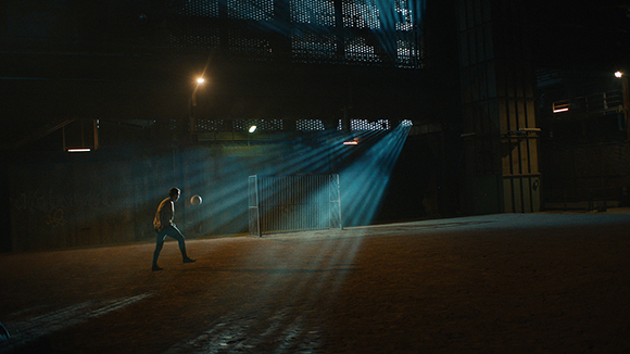 A person kicks a ball in a dark industrial area under bright beams of light.
