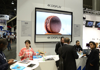 A large screen displays a close-up image of a chocolate macaron at a trade show booth.