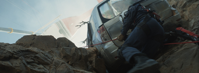 A climber in dark gear works near a silver car stuck on a steep, rocky cliff under a bright sky.