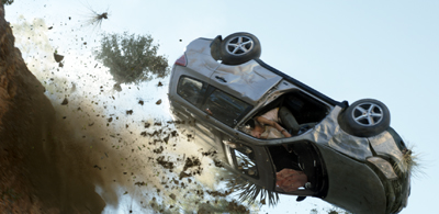 A silver SUV mid-air, flipping upside down near a dirt cliff with debris flying.