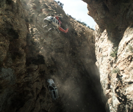 A helicopter flies through a deep, narrow canyon between steep, rocky cliffs.