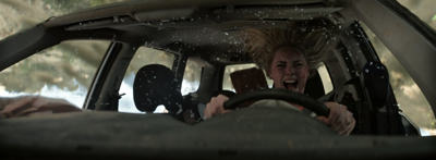 A woman with blonde hair screams in terror while gripping a steering wheel inside a moving vehicle.