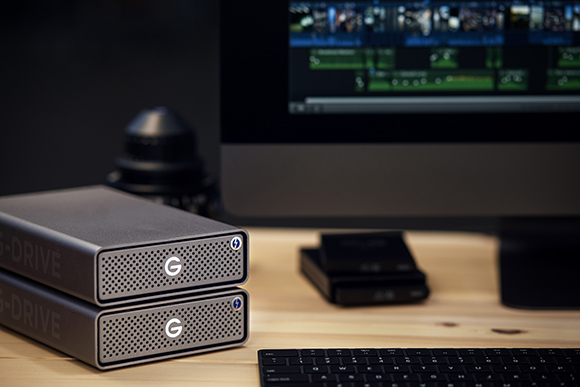 Two silver G-DRIVE external hard drives stacked on a light wooden desk in front of a computer monitor.