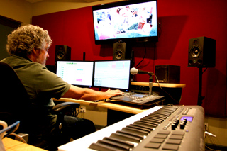 A man sits at a desk with dual monitors, a large screen, studio monitors, and a MIDI keyboard in a red-walled studio.