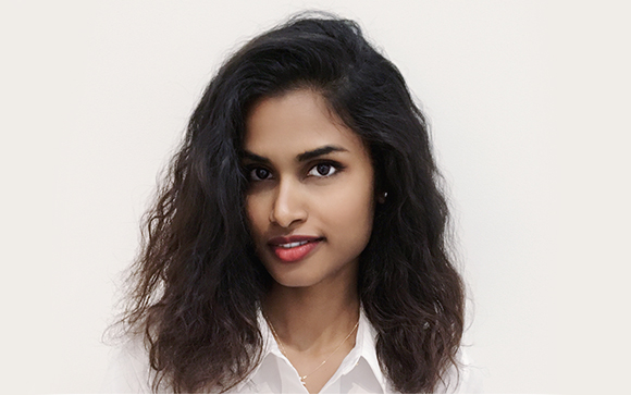 A woman with long, dark wavy hair and a white shirt looks toward the camera against a plain white background.
