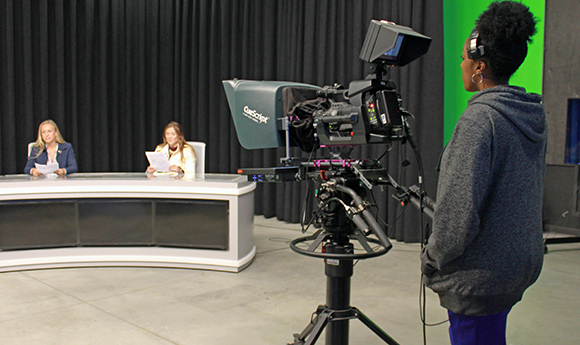 A camera operator films two women seated at a curved white news desk in a studio with black curtains.