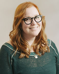 A smiling woman with wavy red hair and black-rimmed glasses wearing a green shirt.