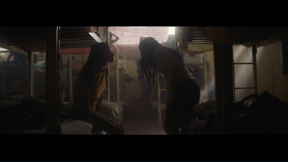 Two people sit on bunk beds in a dark, narrow room with sunlight streaming through a window.